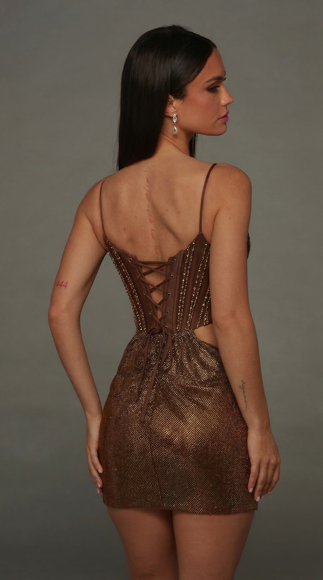 A woman models the Short Homecoming Dress: Alyce 40174 in brown, featuring beaded embellishments. She stands with her back to the camera and her head gently turned to the side against a plain gray background.